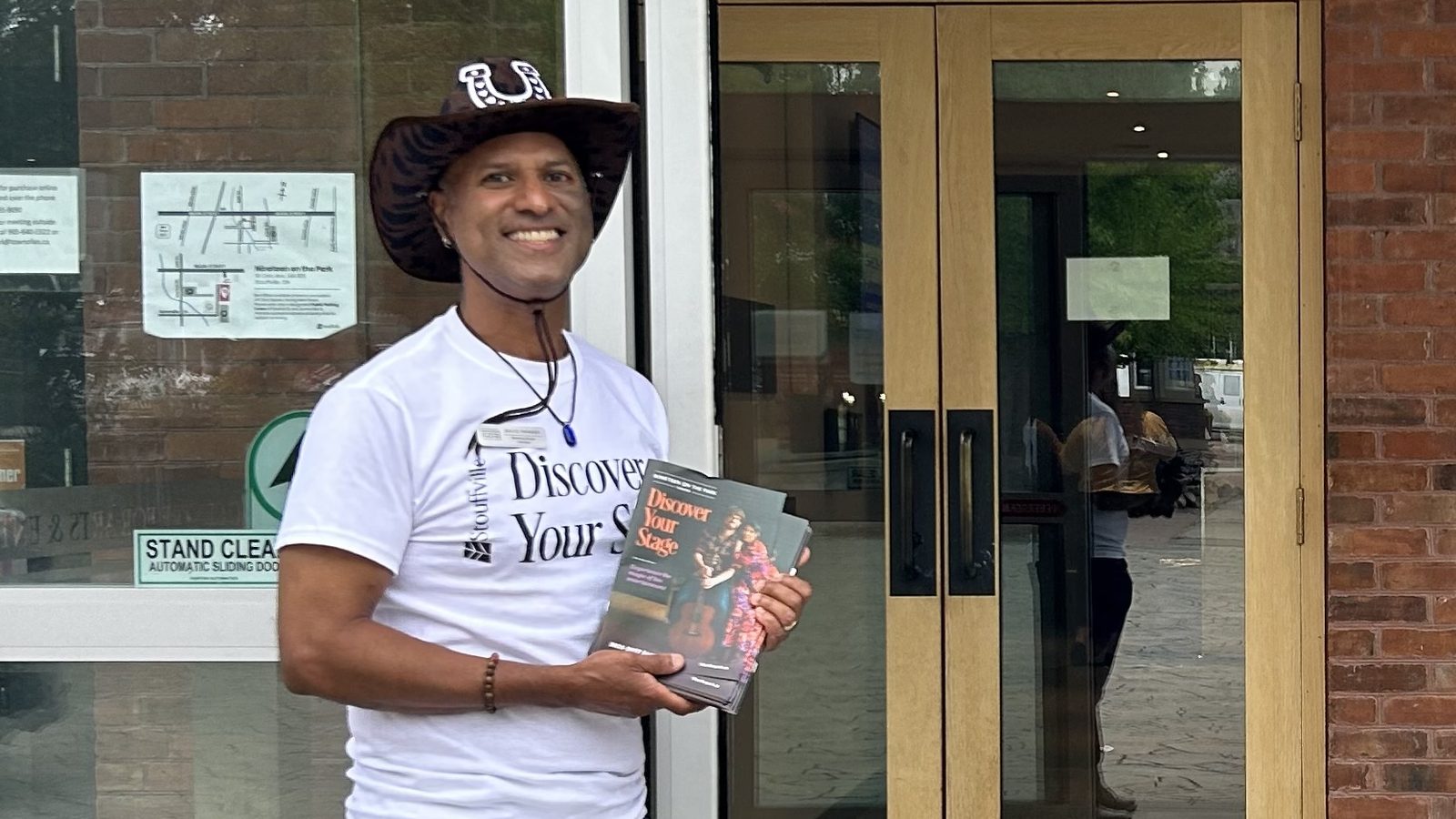 David Parmer volunteering outside the doors of 19 on the Park, wearing a Discover Your Stage volunteer shirt and holding a stack of season programmes