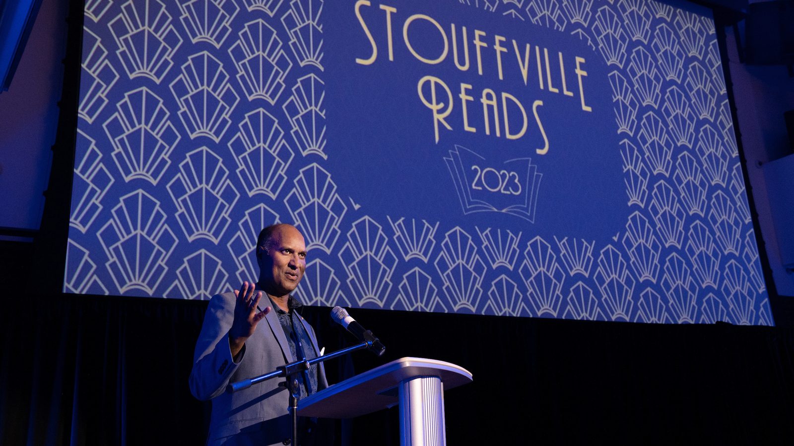 David Parmer speaking at a podium at the Stouffville Reads 2023 event, the illustrated event title projected on the screen behind him